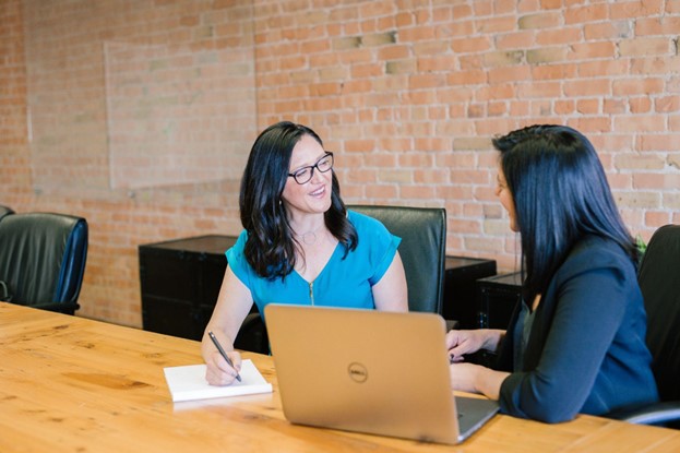 Interior, two women working together at a table with a laptop and notepad. Investing